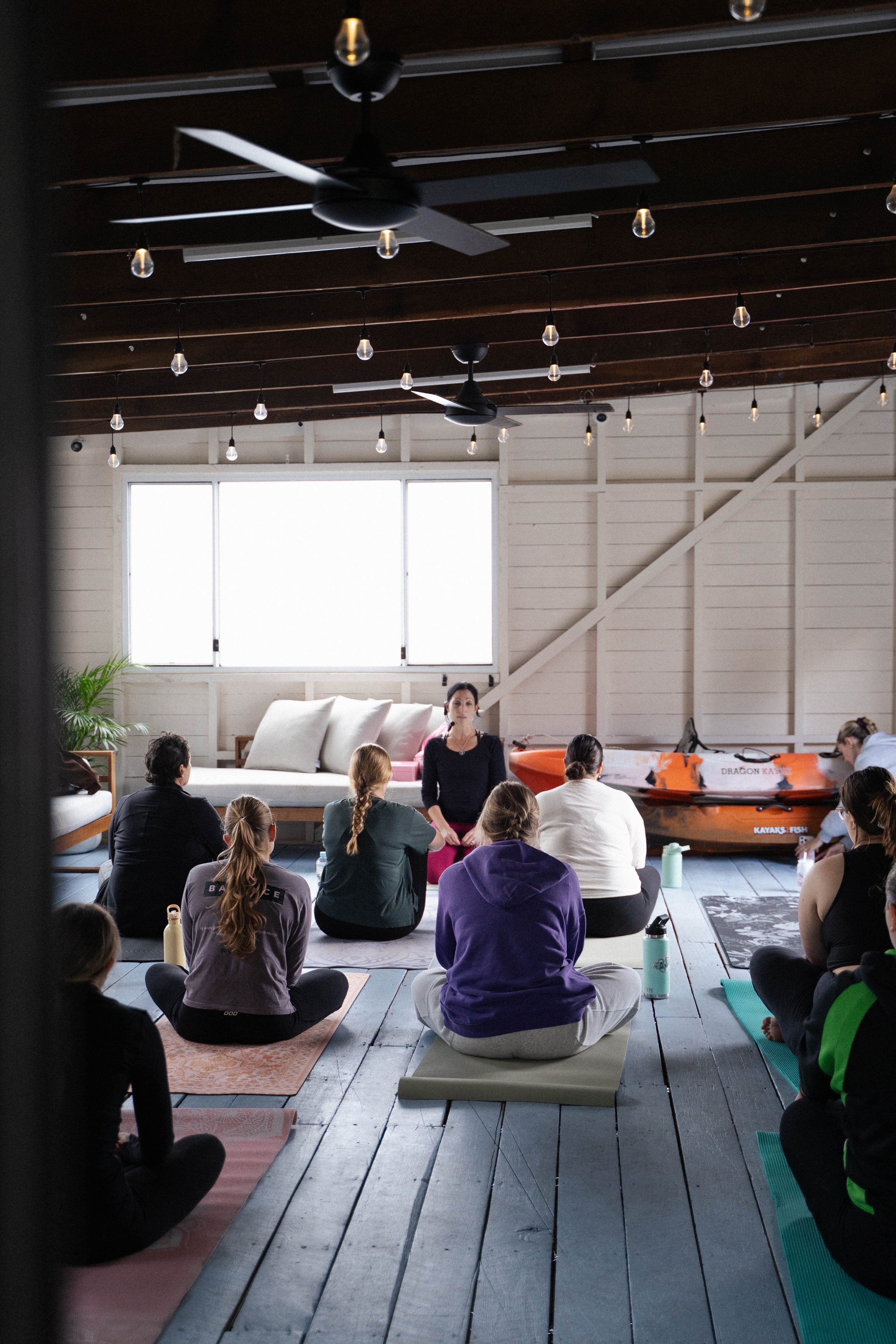 Women seated on yoga mats during a guided indoor yoga class in a bright studio with timber floors, creating a calm and welcoming environment at a The Girls Who Gather wellness event.