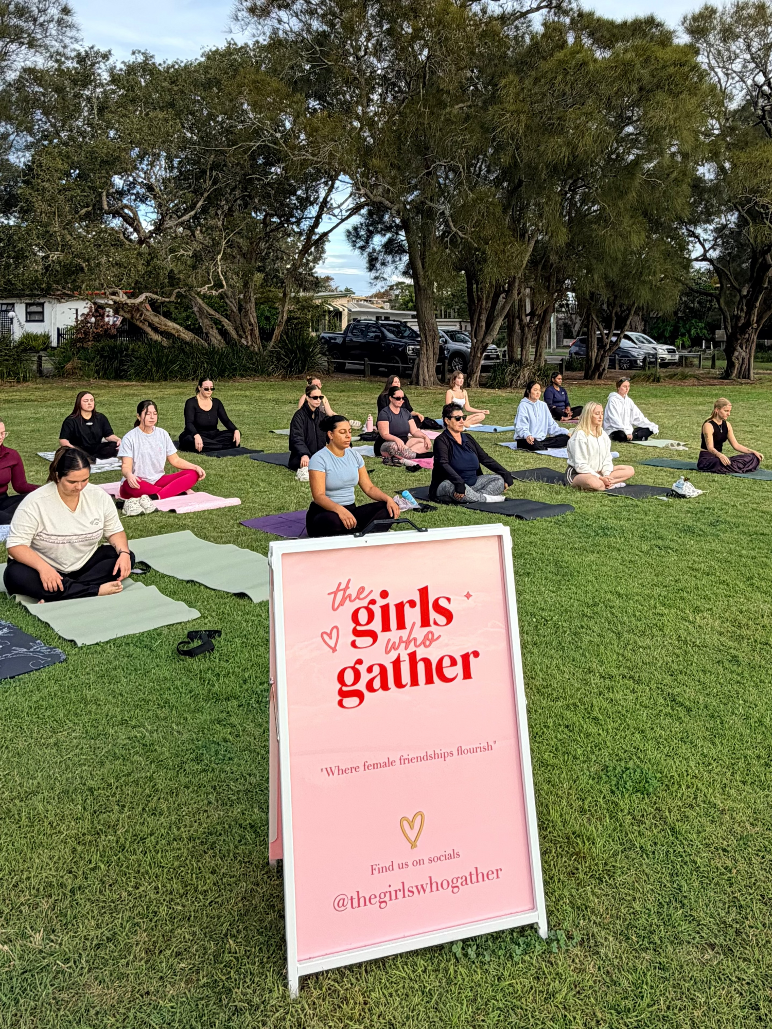 Women seated on yoga mats during an outdoor yoga session on a grassy park, with a The Girls Who Gather welcome sign in the foreground, creating a peaceful and welcoming community wellness event.