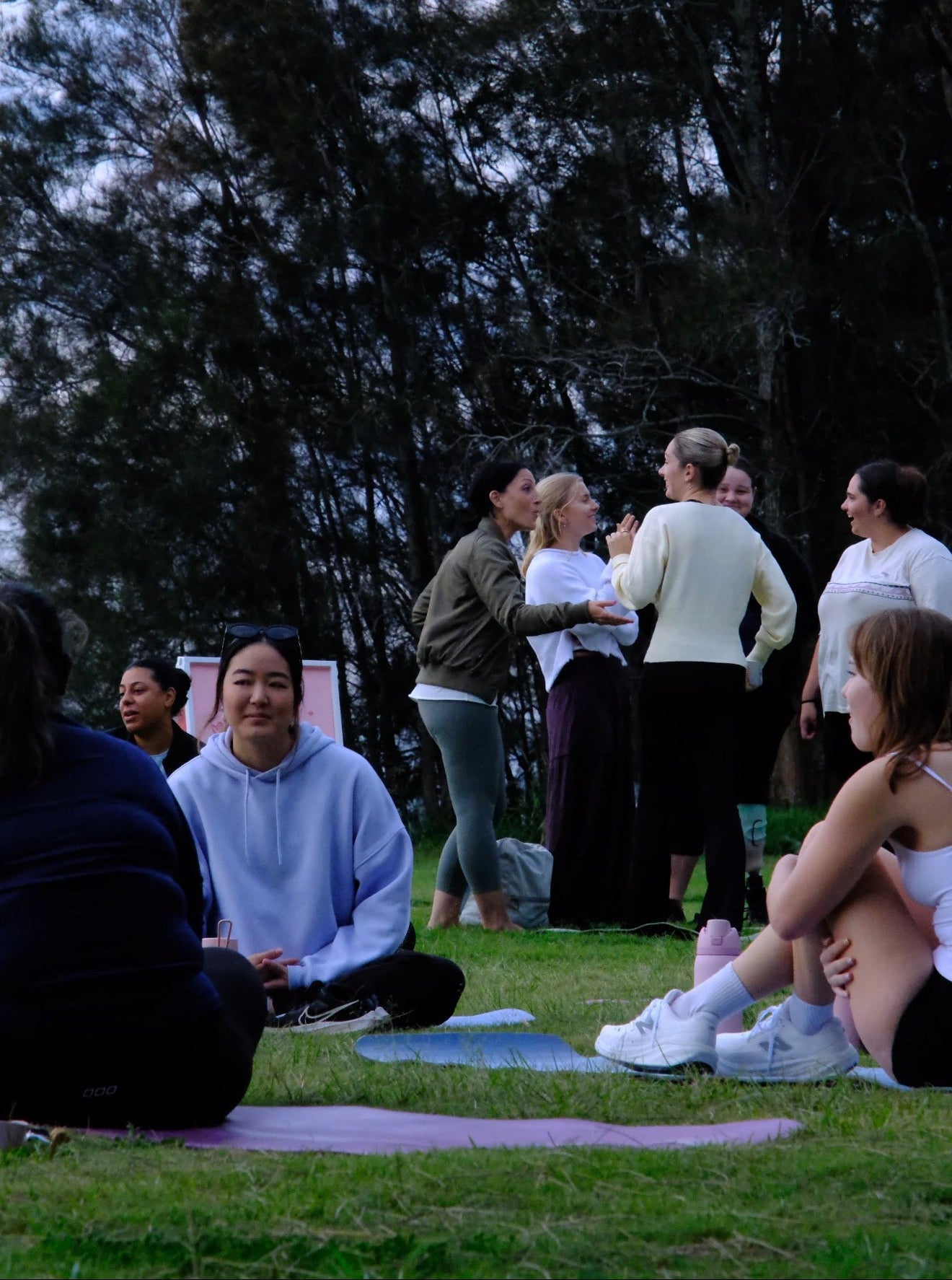 Women relaxing and chatting on yoga mats after an outdoor yoga session in a park, capturing the friendly and welcoming atmosphere of a The Girls Who Gather community wellness event.