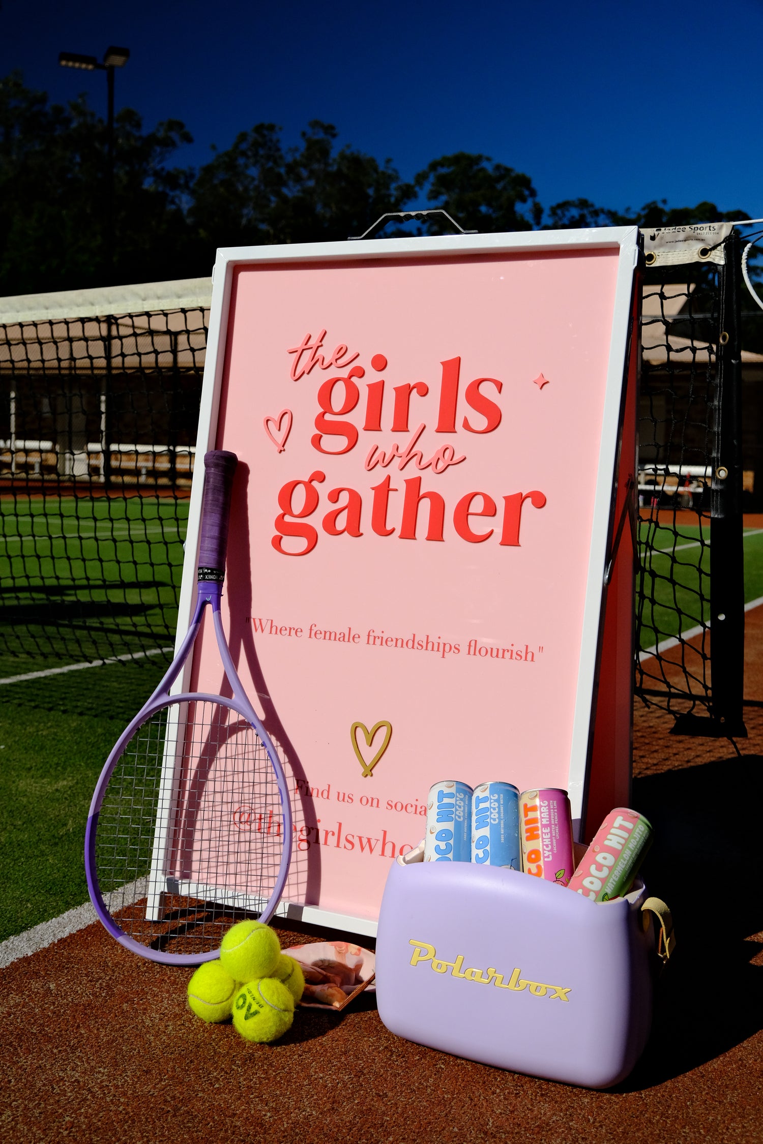 The Girls Who Gather event display on a tennis court featuring a pink welcome sign, a purple tennis racquet, tennis balls, Polarbox cooler bag, and Coco Hit sparkling water cans arranged for a community tennis event.