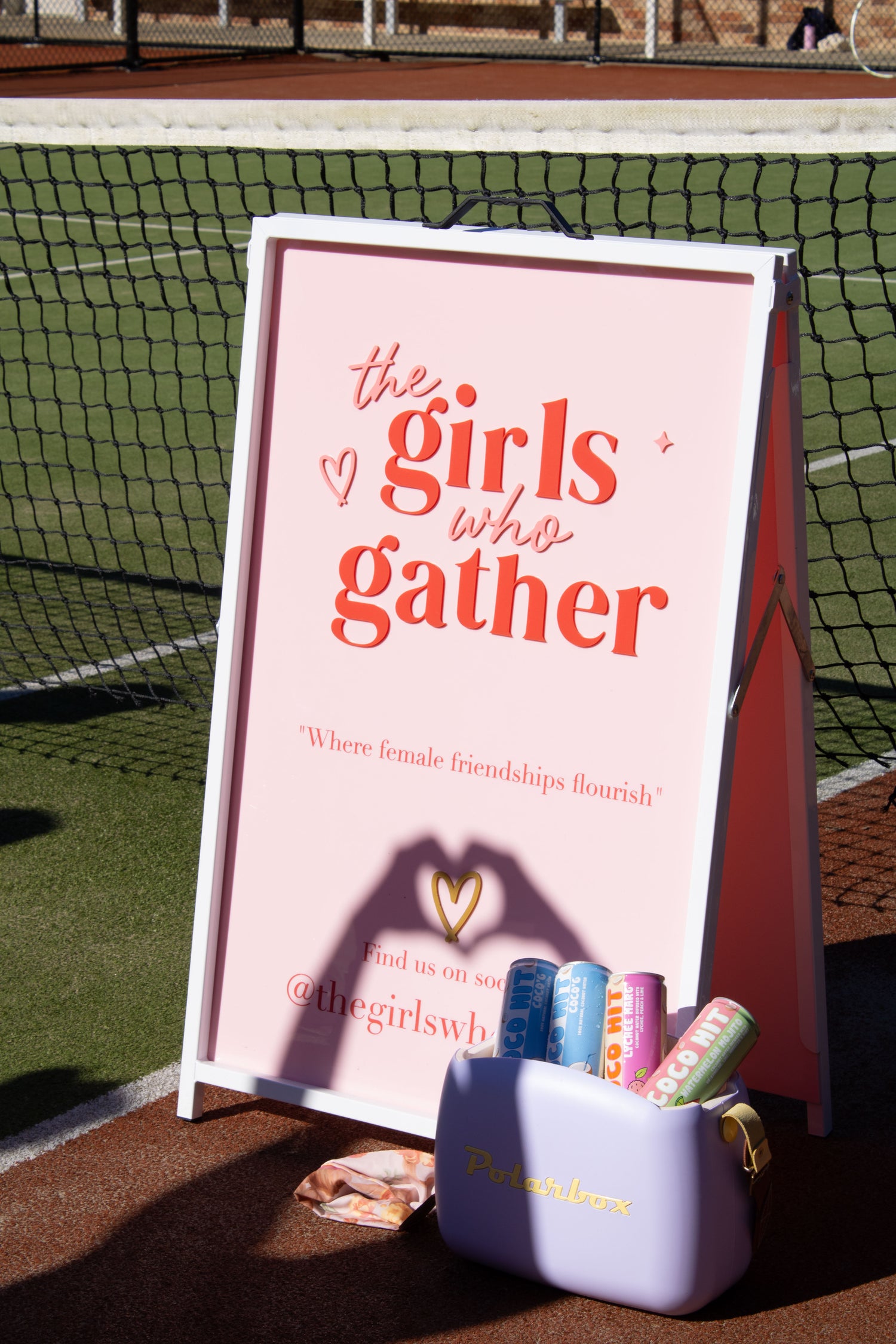 The Girls Who Gather welcome sign displayed beside a tennis court with sponsor products and giveaway items, ready for a women's social tennis event on the Central Coast.