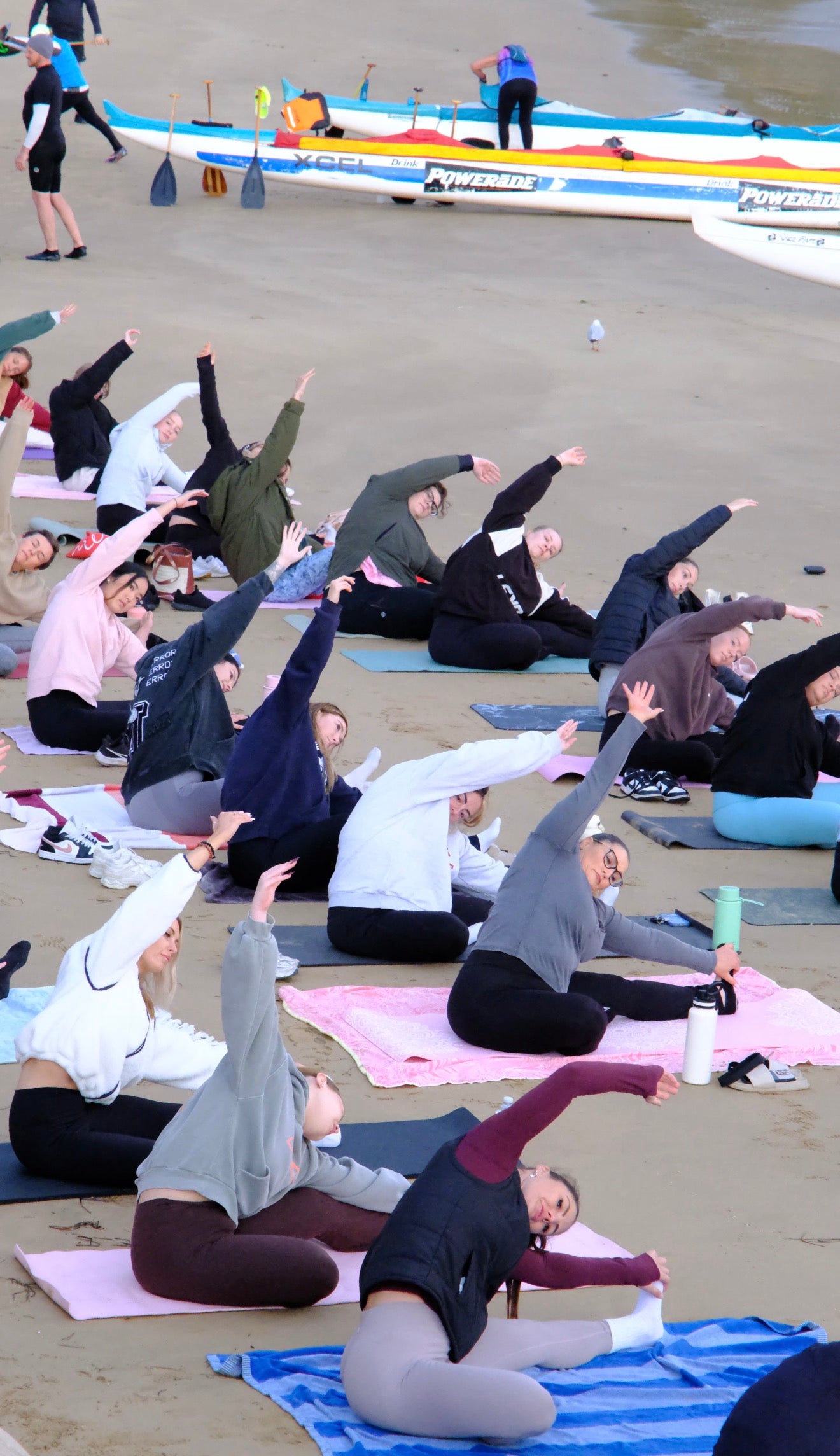 Women taking part in a beach yoga session during a The Girls Who Gather wellness event on the Central Coast, stretching together on yoga mats with paddle boats and the ocean in the background.