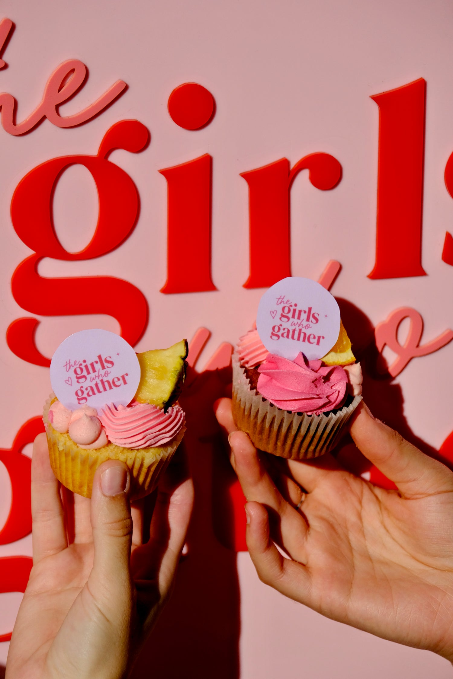 Close-up of two branded cupcakes featuring The Girls Who Gather logo, held by attendees at a women's community event on the Central Coast. Custom desserts created for a TGWG event celebrating connection, friendship and local businesses.
