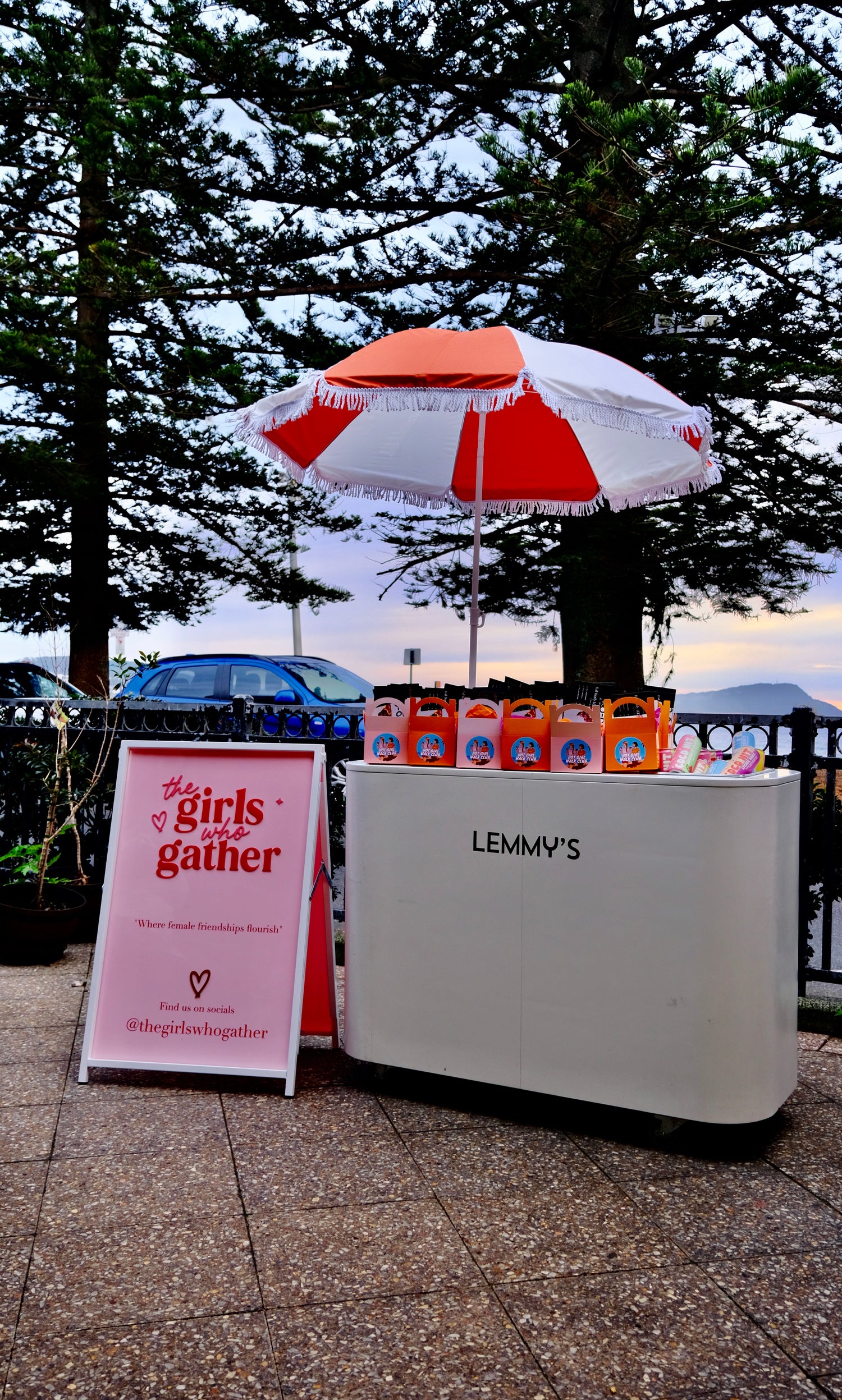 The Girls Who Gather Hot Girl Walk Club event setup outside LEMMY'S in Terrigal, featuring a branded welcome sign, community table, goodie bags and beach umbrella before a free women's community walk on the Central Coast.